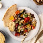 A white plate with roasted vegetables, pita bread, hummus, crumbled feta, and olives, placed on a marble surface with a fork and a small bowl of hummus nearby.
