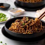 A black bowl filled with stir-fried noodles and beef, garnished with chopped green onions, with chopsticks lifting some noodles. Other small dishes are in the background.
