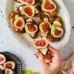 A hand holds a piece of toasted bread topped with cheese and fresh fig, with more similar brie and fig crostini arranged on a white platter.