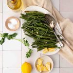 Oval platter of cooked green beans with serving utensils, surrounded by lemon wedges, salt, parsley, and a bottle of oil on a tiled surface.