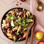 A salad with sliced apples, arugula, pomegranate seeds, pecans, dried cranberries, and crumbled cheese in a black bowl with wooden salad servers.
