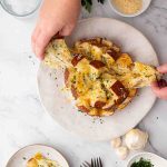 Hands pulling apart a round loaf of bread filled with melted cheese and herbs on a white plate, with ingredients like garlic and parmesan nearby.