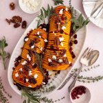 Sliced roasted butternut squash garnished with pecans, goat cheese, pomegranate seeds, fresh herbs, and served on an oval white platter with utensils.