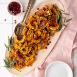 Oval platter with roasted delicata squash slices, garnished with pomegranate seeds, herbs, and pistachios. A serving spoon, bowl, and fresh sage are also visible.
