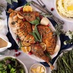 Sliced roasted turkey with herbs, lemon wedges, and onions on a platter, surrounded by mashed potatoes, green beans, salad, and gravy on a table.