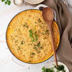 A large round casserole dish filled with baked mac and cheese topped with breadcrumbs and garnished with parsley, with a wooden spoon resting on top.