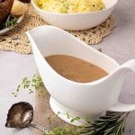 A white gravy boat filled with brown gravy sits on a beige cloth next to a spoon, with mashed potatoes and herbs in the background.