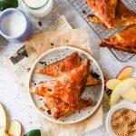 Three golden-brown apple turnovers on a white plate, surrounded by apple slices, a cup of milk, a bowl of sugar, and additional turnovers on a cooling rack.