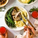 A plate of mashed potatoes, green beans, and beef with mushroom sauce, next to two glasses of red drink. A hand is holding a fork and knife, cutting into the food.