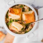 Individual serving of chicken pot pie topped with puff pastry cubes in a white ramekin, with a spoon and a sprig of thyme.