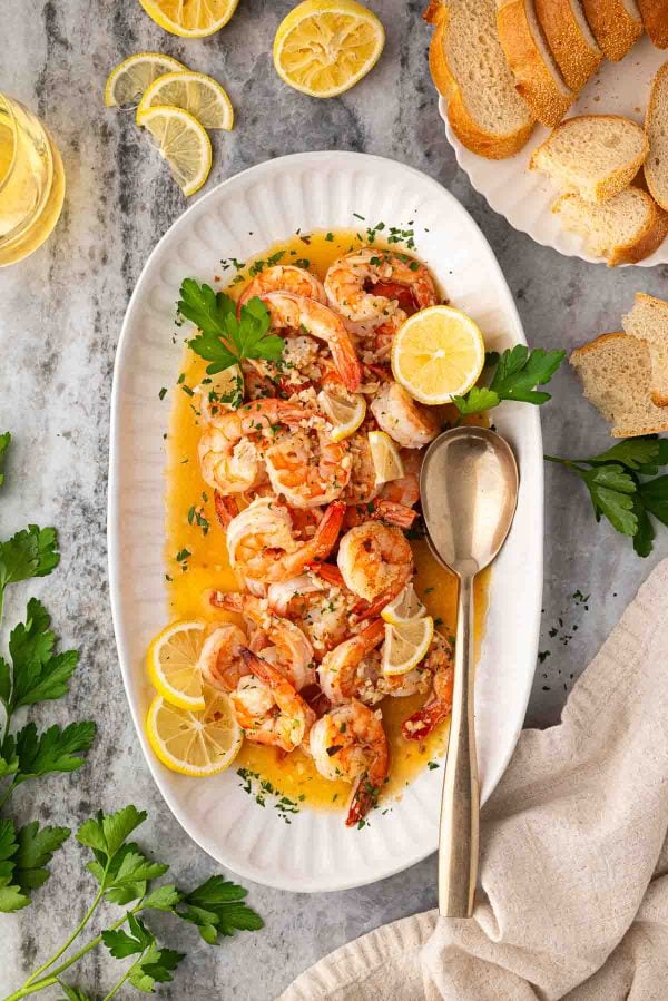 A white oval platter with cooked shrimp in a buttery sauce, garnished with lemon slices and parsley, alongside a serving spoon, bread slices, and a glass of white wine.