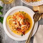 A white plate with shrimp in a garlic butter sauce, garnished with lemon slices and parsley, next to slices of bread and a fork and spoon.