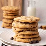A stack of bacon chocolate chip cookies on a white plate with chocolate chips scattered nearby and milk in the background.