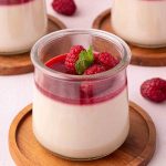 A glass jar of panna cotta topped with raspberry sauce, fresh raspberries, and a mint leaf, placed on a wooden coaster.