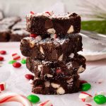 A stack of four christmas brownies with white chocolate chips and crushed peppermint on top, surrounded by candy pieces and broken peppermint sticks.