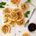 A pile of savory pastry pinwheels garnished with herbs on a white surface, next to a knife, parsley sprigs, and a small bowl of dark dipping sauce.