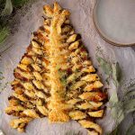 Puff pastry twisted and arranged in the shape of a Christmas tree, topped with melted cheese, surrounded by herbs, with stacked plates nearby.