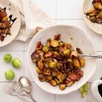 A bowl of roasted Brussels sprouts with bacon sits on a white tiled surface, surrounded by scattered Brussels sprouts, serving utensils, and two small plates with servings.