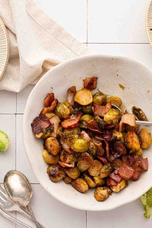 A bowl of roasted Brussels sprouts with bacon sits on a white tiled surface, surrounded by scattered Brussels sprouts, serving utensils, and two small plates with servings.