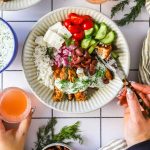 A bowl with rice, grilled chicken, tzatziki, sliced cucumber, tomato, feta, red onion, and olives. A person is holding a fork and another hand holds a glass with a pink drink.