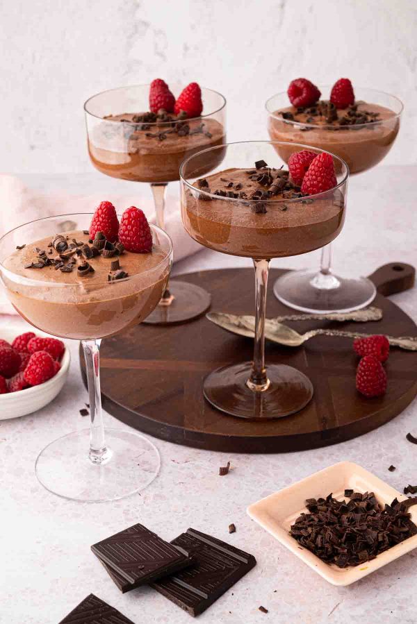 Four glass cups of chocolate mousse topped with raspberries and chocolate shavings are displayed on a round wooden tray with extra raspberries and chocolate pieces nearby.