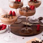 Four coupe glasses filled with chocolate mousse, topped with raspberries and chocolate shavings, are placed on a round wooden tray, with extra raspberries and chocolate nearby.