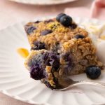 A slice of baked oatmeal with blueberries on a white plate, garnished with more blueberries and a dollop of cream, with a fork holding a piece.