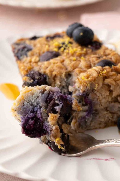A slice of baked oatmeal with blueberries on a white plate, garnished with more blueberries and a dollop of cream, with a fork holding a piece.