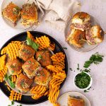 A plate of small sandwiches topped with herbs is served with waffle fries. Additional sandwiches and fries are on side plates, garnished with parsley.