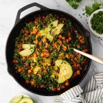 A cast-iron skillet with a cooked mixture of ground meat, sweet potatoes, greens, melted cheese, and sliced avocado, garnished with chopped herbs.