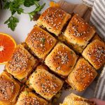 A hand holding a Hawaiian roll breakfast slider topped with seasoning; several sliders are arranged on a tray with herbs and an orange slice nearby.