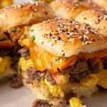 Close-up of breakfast sliders with scrambled eggs, sausage, cheddar cheese, and bacon on sesame seed buns, arranged on a tray. Text reads, "How to Make Easy Breakfast Sliders.