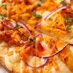 Flatbread topped with melted cheese, shredded buffalo chicken, red onion slices, green herbs, and a drizzle of buffalo sauce, with part of the flatbread cut into slices.