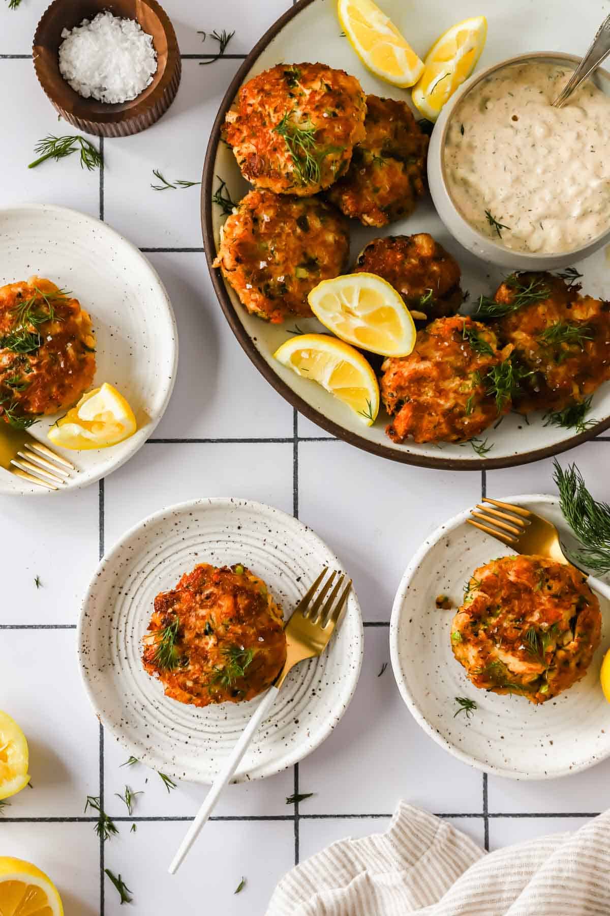 Top-down view of plates with crab cakes garnished with lemon wedges and fresh herbs, served with a bowl of dipping sauce and a small dish of salt.