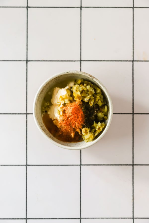 A small bowl containing chopped pickles, mayonnaise, and spices sits on a white tile surface with a black grid pattern.