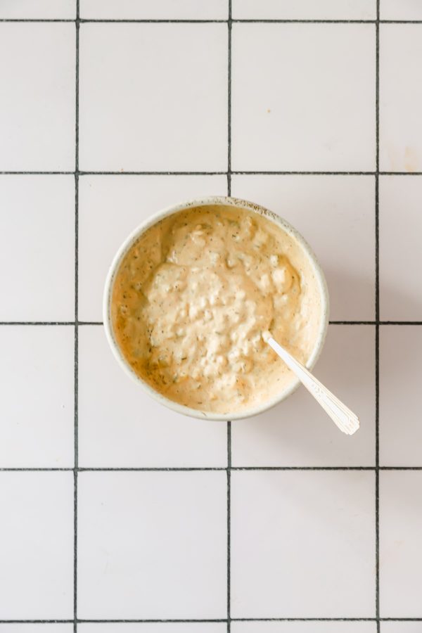 A bowl of creamy dip with herbs and a spoon, placed on a white tiled surface with black grout lines.