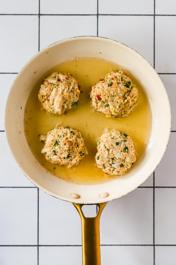 Four uncooked crab cakes are arranged in a white frying pan with oil, set on a white tiled surface.