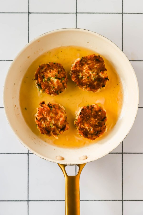 A white frying pan with four golden-brown patties frying in oil on a white tiled surface.