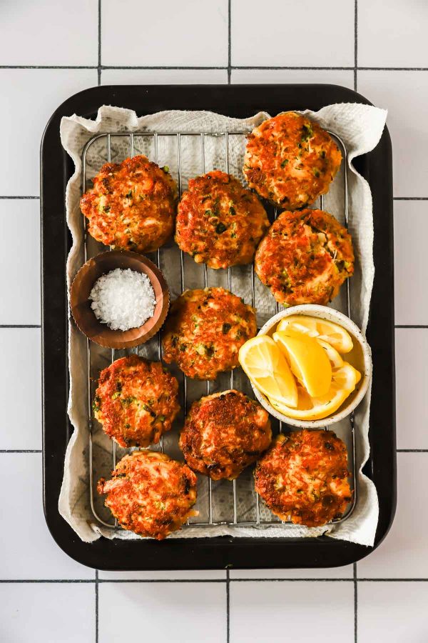 A tray of golden-brown crab cakes on a wire rack, served with a bowl of coarse salt and a bowl of lemon wedges, on a parchment-lined baking sheet.
