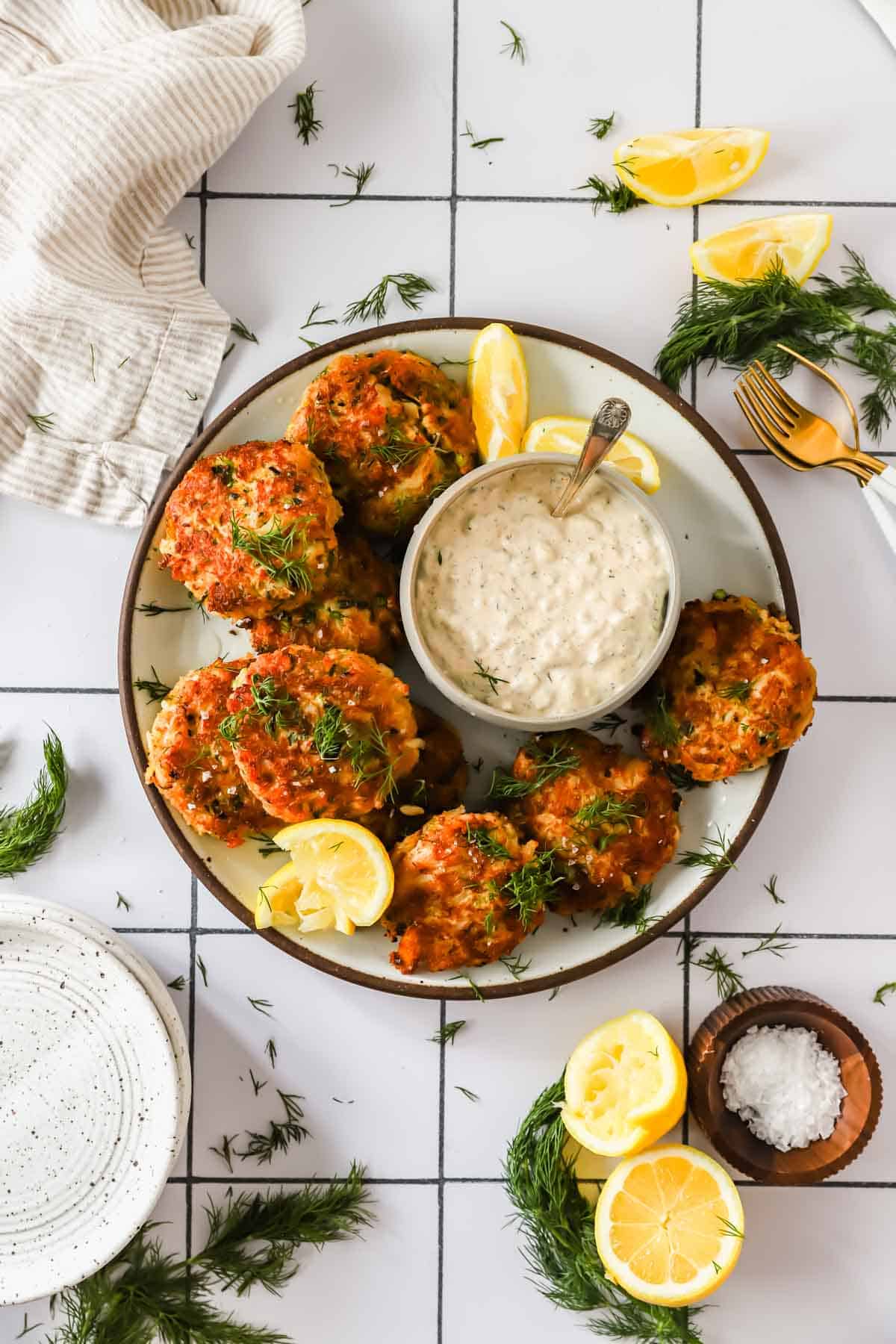 A plate of crab cakes garnished with fresh dill and lemon wedges, served with a bowl of creamy dipping sauce on a tiled white table.