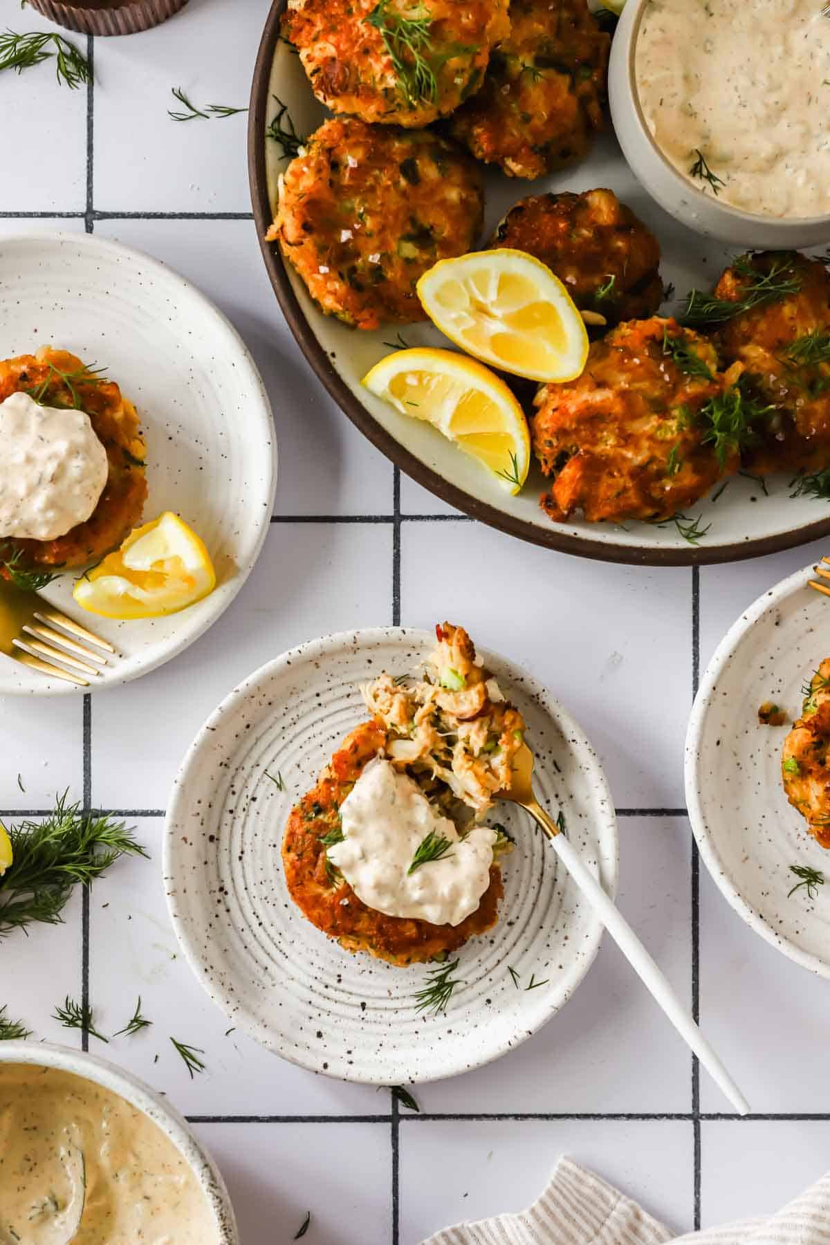 Plates of crab cakes topped with creamy sauce, garnished with fresh dill and lemon wedges, arranged on a white tiled surface.