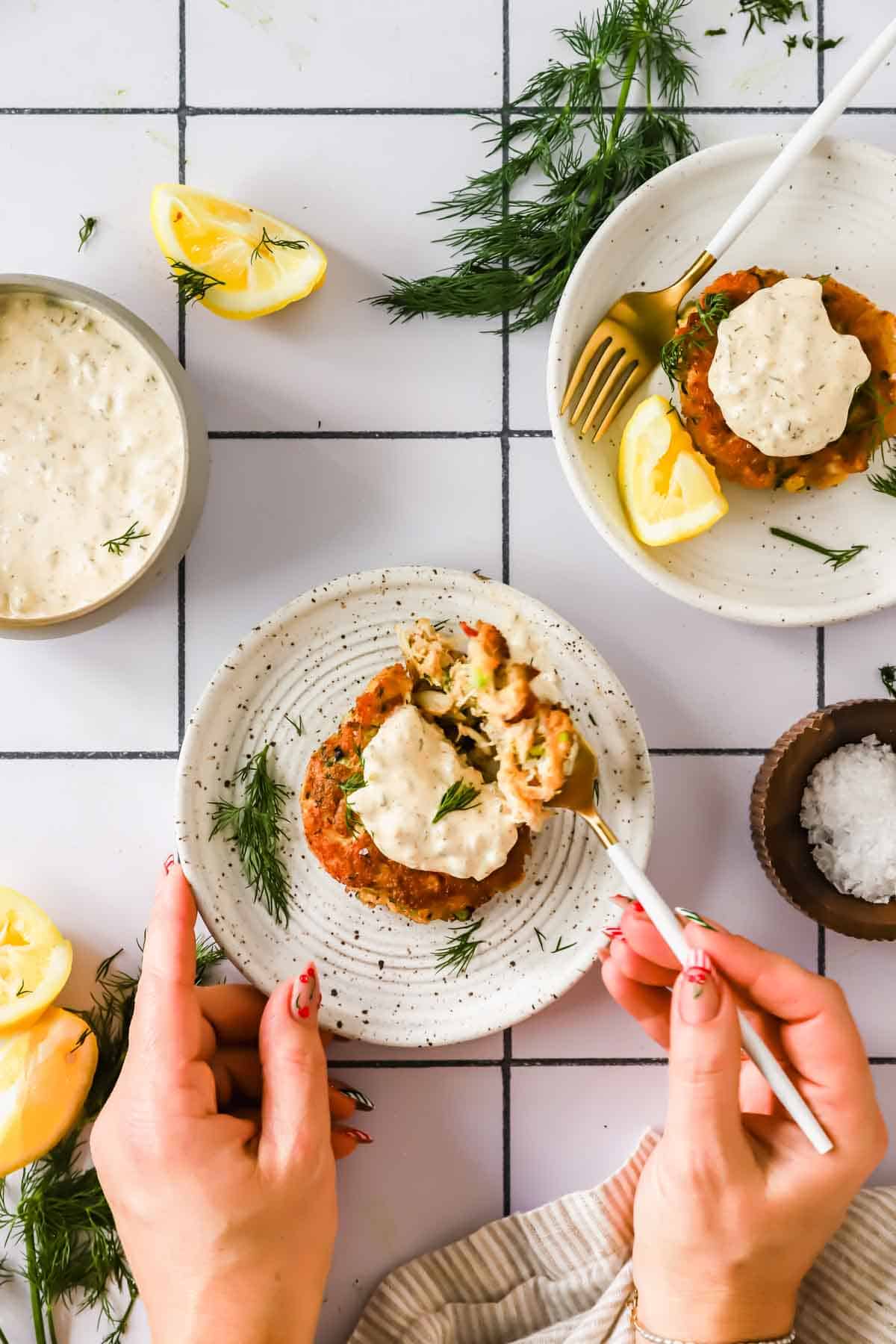 Two plates with salmon patties topped with creamy sauce, garnished with dill and lemon wedges, on a tiled table. A hand holds a fork with a bite, and sauces are nearby.