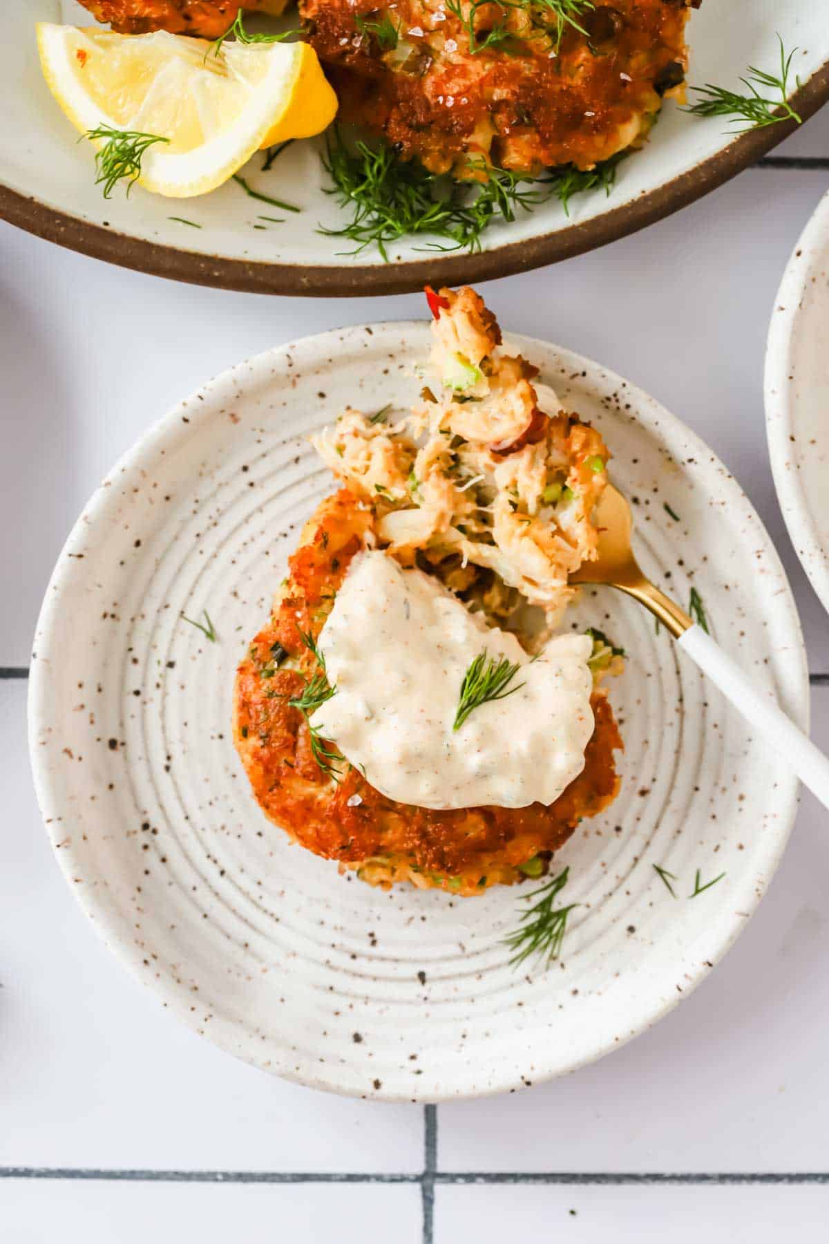 A crab cake topped with tartar sauce and a sprig of dill on a speckled plate, with a fork holding a bite-sized piece.