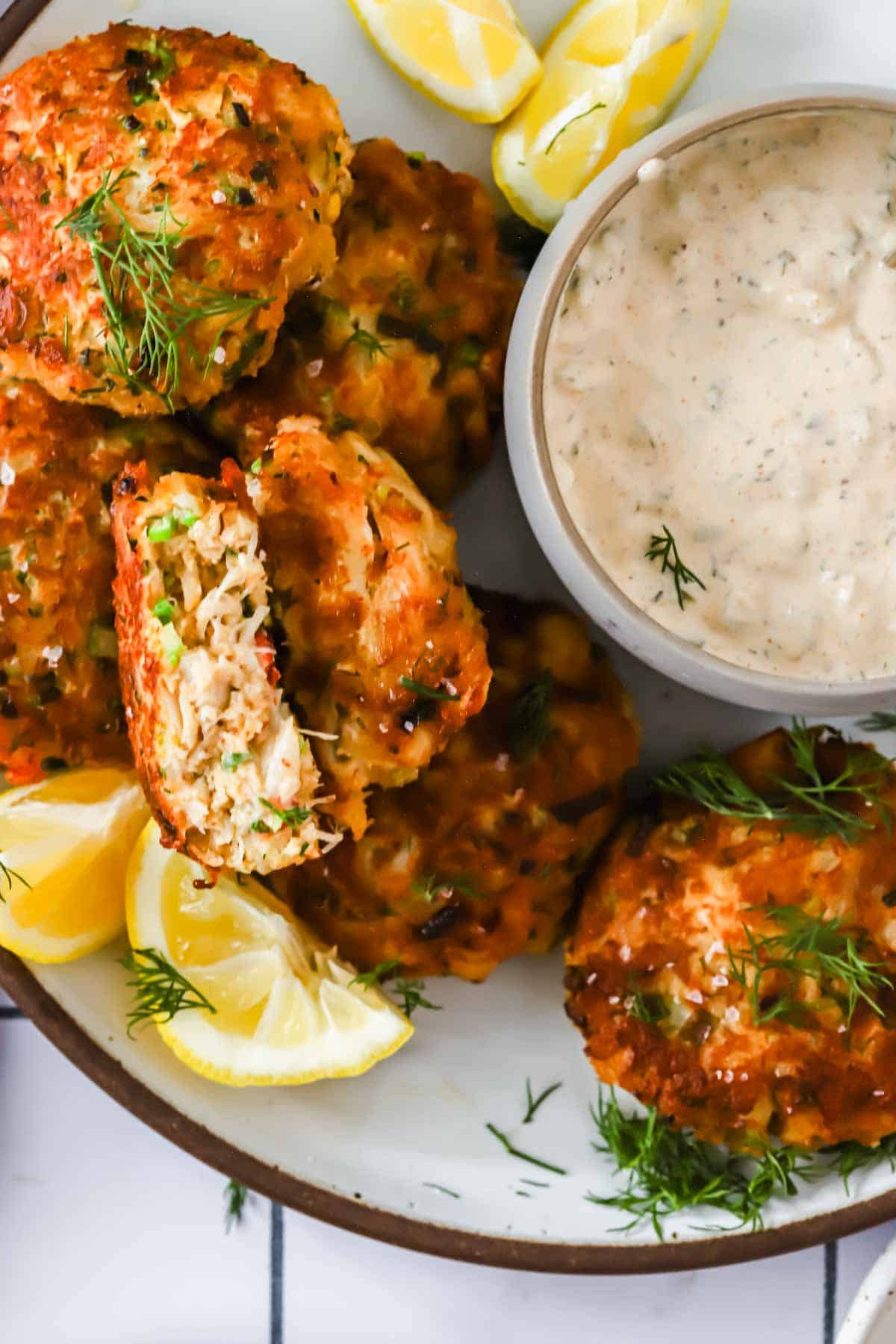 A plate of crab cakes garnished with fresh dill and lemon wedges, served with a bowl of creamy dipping sauce.