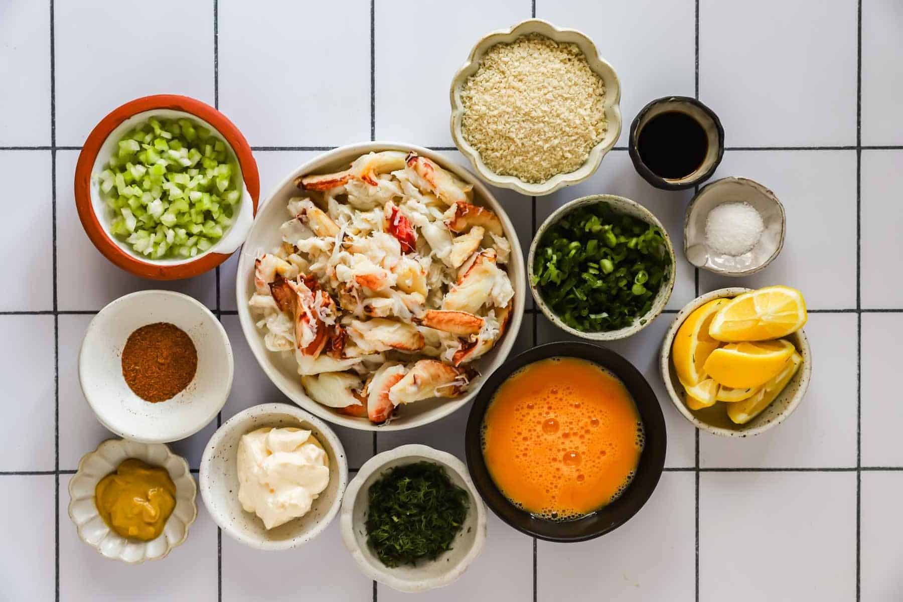 A variety of ingredients in separate bowls, including crab meat, chopped vegetables, eggs, seasonings, lemon wedges, breadcrumbs, and sauces, arranged on a white tiled surface.