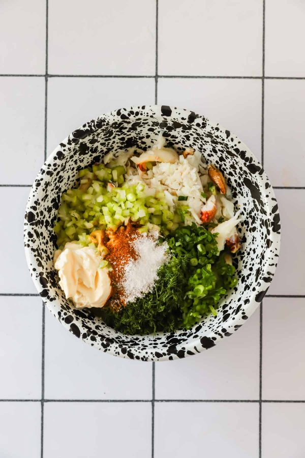 A bowl containing chopped celery, crab meat, mayonnaise, green onions, dill, spices, and salt on a white tiled surface.