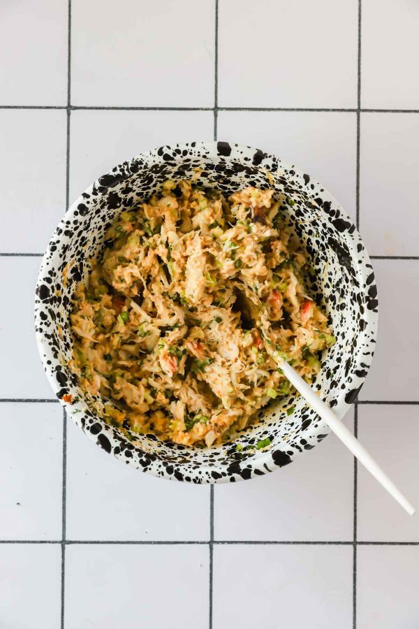A speckled bowl containing a mixed filling with chopped vegetables and herbs sits on a white tiled surface with a white spoon inside.