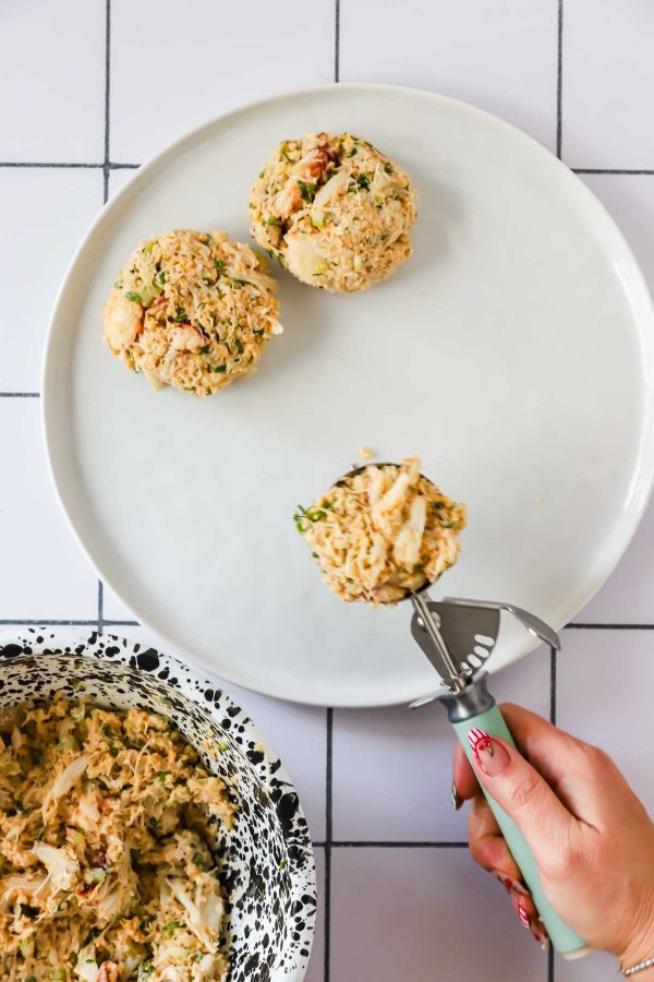 A hand uses an ice cream scoop to portion a mixture onto a white plate, with three formed scoops and a bowl of the same mixture nearby on a tiled surface.