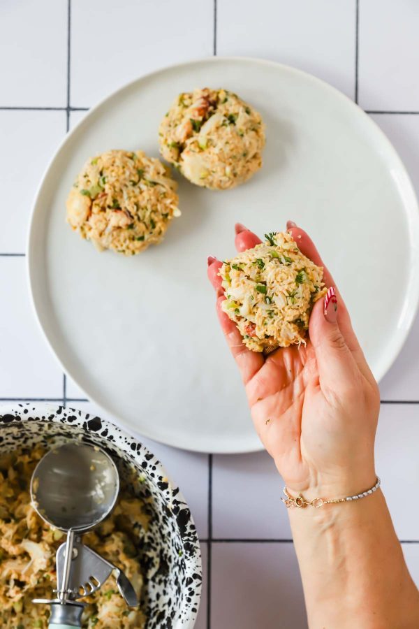 A hand forms a round patty from a mixture, with two more patties on a plate and a bowl with a metal scoop nearby on a tiled surface.