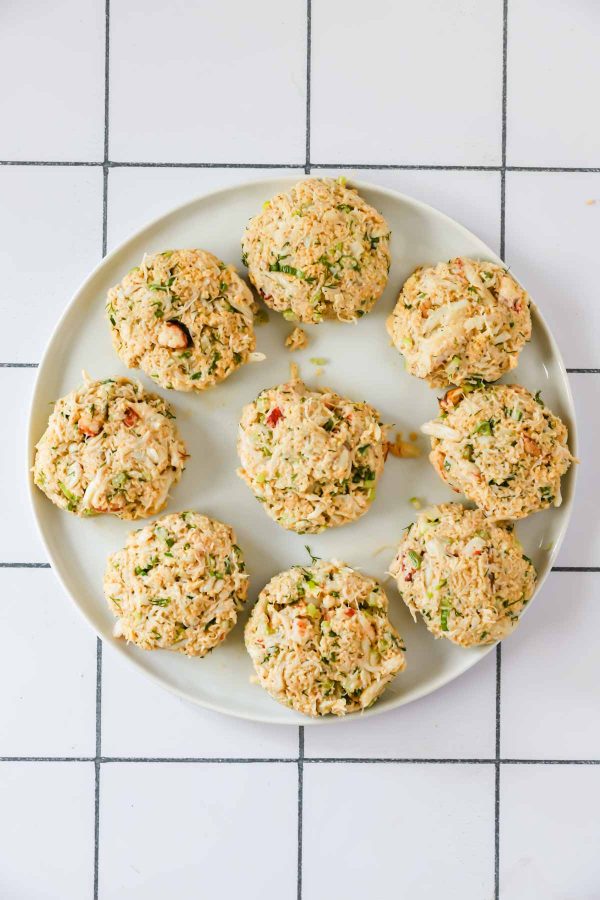 A round white plate holds nine uncooked crab cakes arranged in a circle, sitting on a white tiled surface.